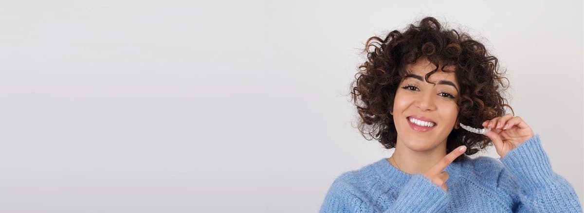 Invisalign-before-and-after-showing-a-smiling-woman-in-a-blue-sweater-holding-and-pointing-to-an-invisalign-tray-against-a-light-background