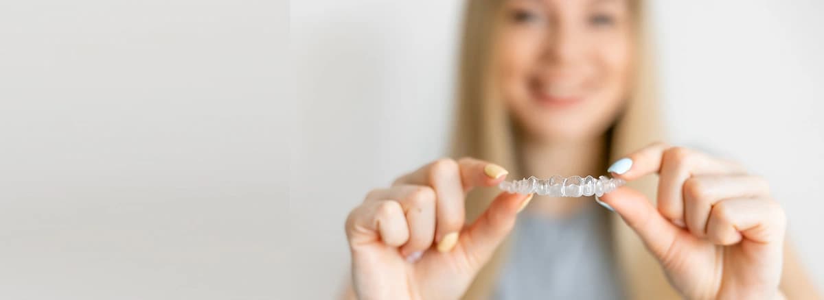 Smiling-woman-holding-clear-aligners-between-her-fingers-showing-invisalign-benefits-blurred-background-with-neutral-lighting-and-focus-on-transparent-teeth-straightening-tray