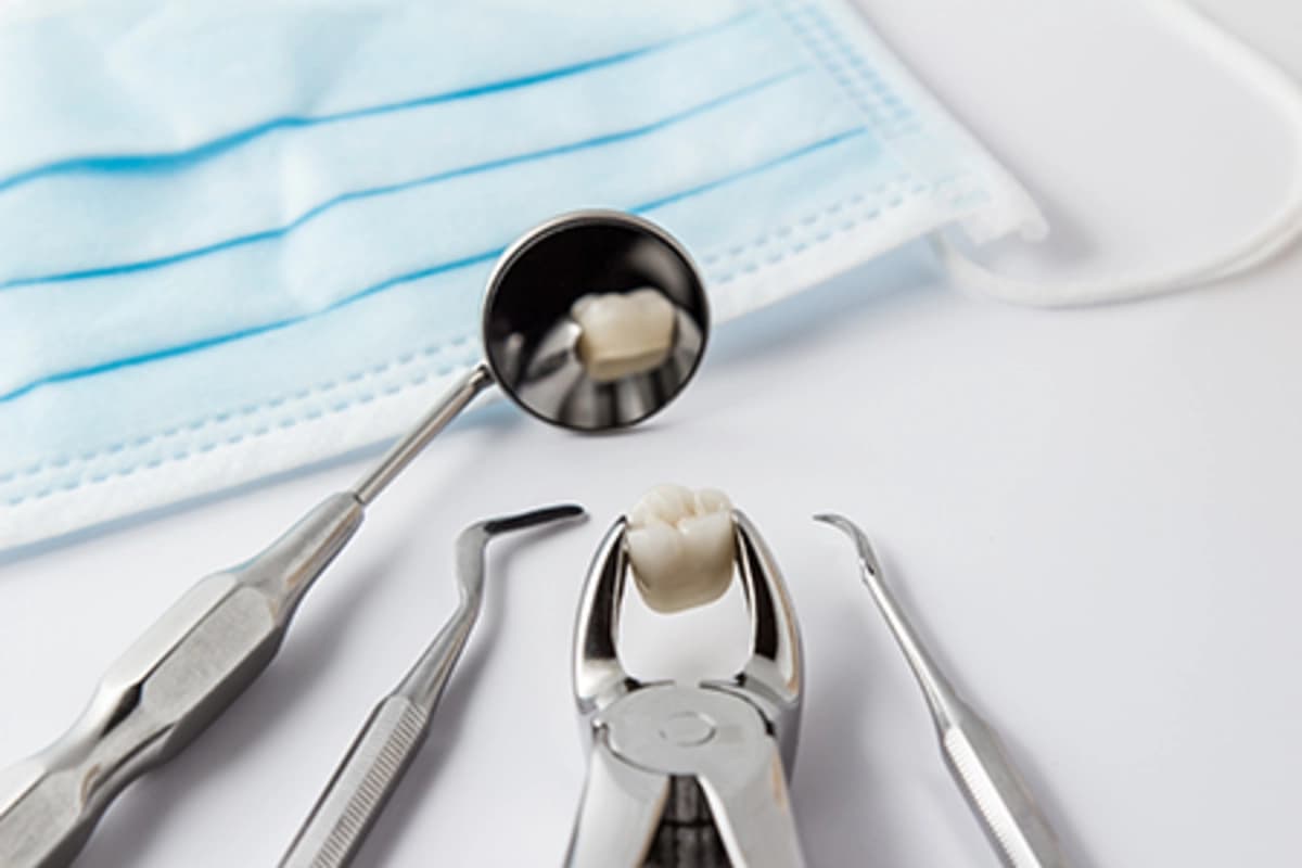 Dental tools and forceps holding extracted tooth on white surface with dental mirror reflecting tooth and blue surgical mask in background