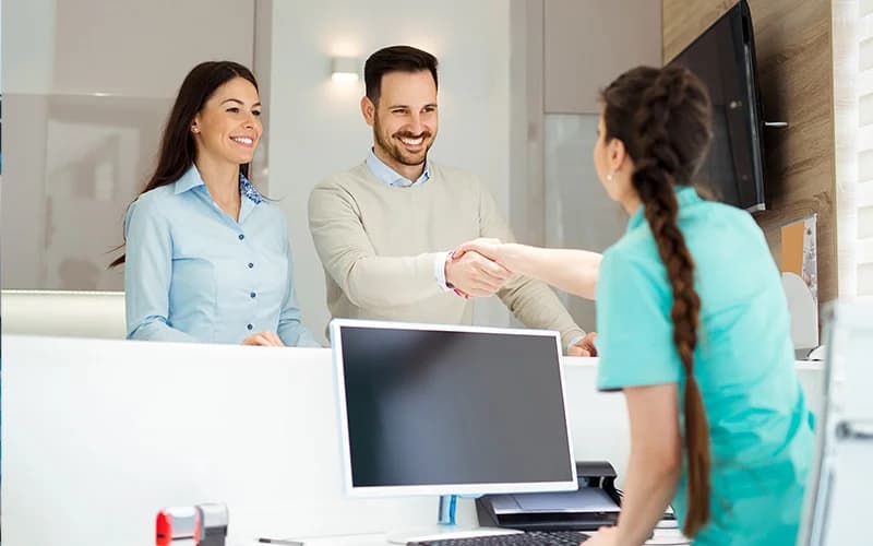 Man and woman smiling at dental receptionist shaking hands in modern office discussing ceramic crowns queens treatment options