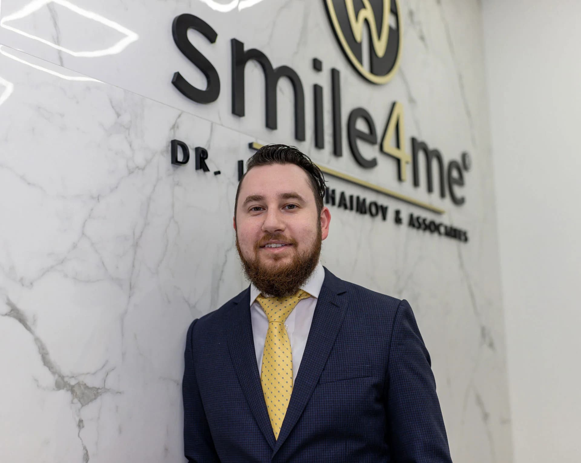 Man in a navy suit and yellow tie standing in front of a marble wall with the smile4me dental care logo