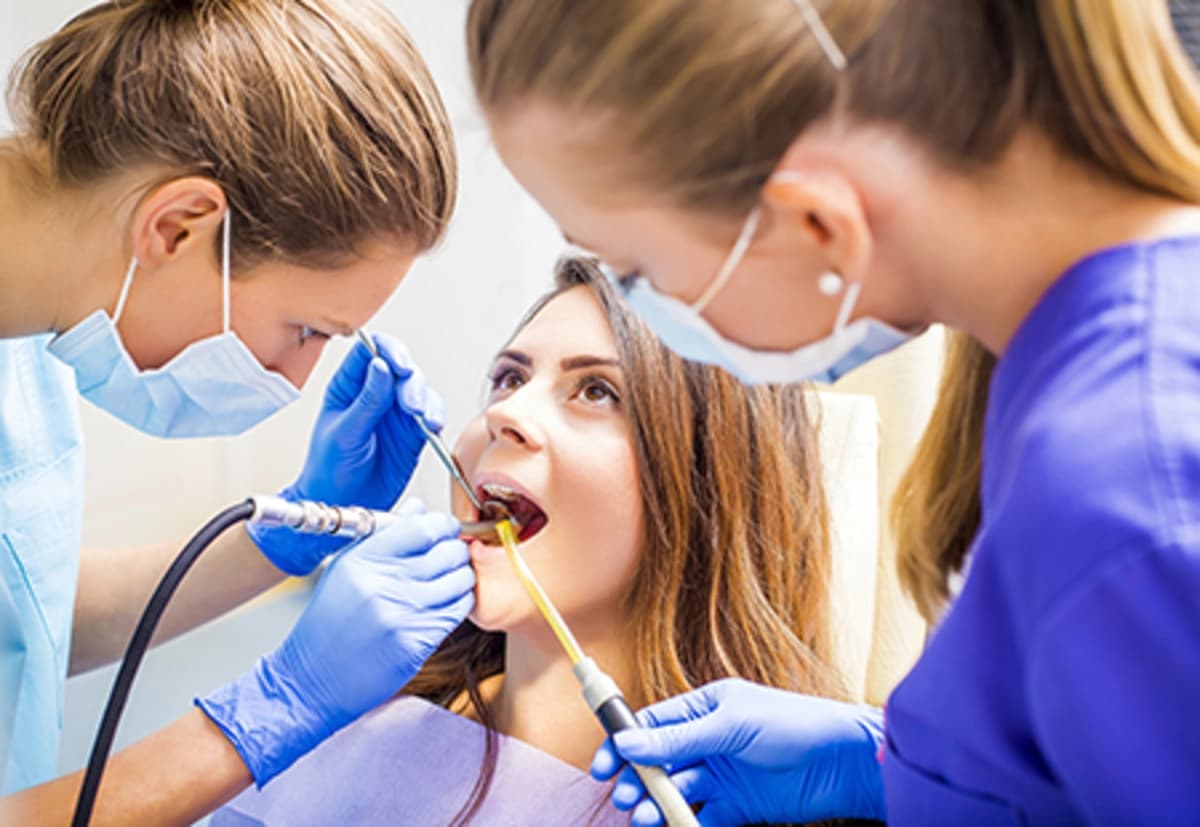 Patient receiving dental cleaning procedure with two dental professionals wearing masks and gloves using tools in a clinical setting