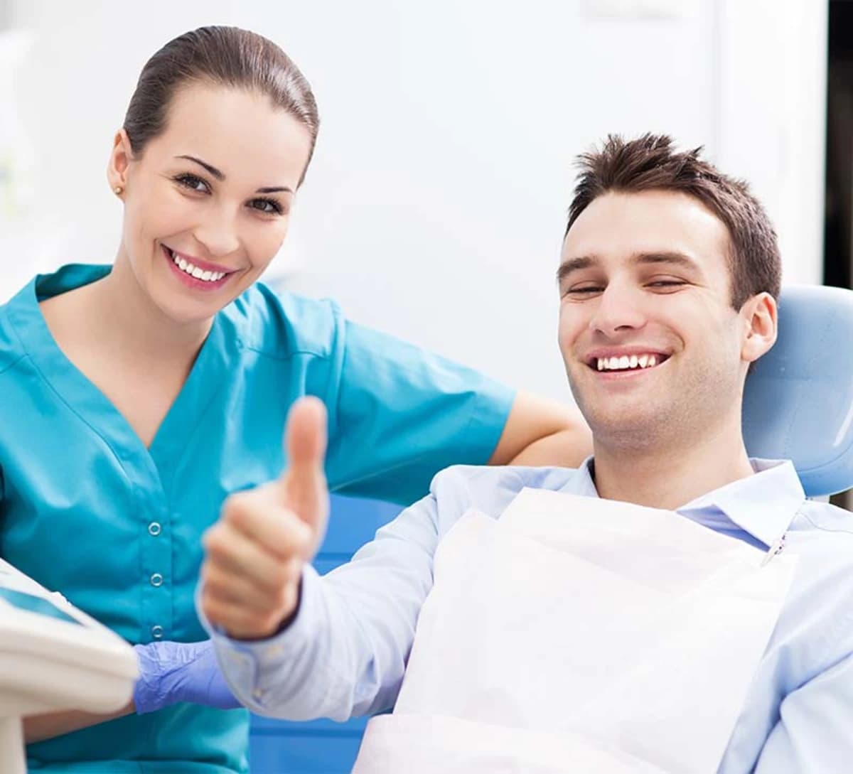 Smiling dentist in blue scrubs and happy male patient in dental chair giving thumbs up wearing bib in clean dental office setting