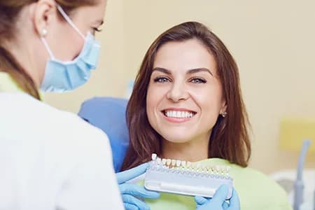 Smiling woman sitting in dental chair while dentist holds tooth shade guide demonstrating options for veneers Astoria patient wants to achieve a brighter smile.