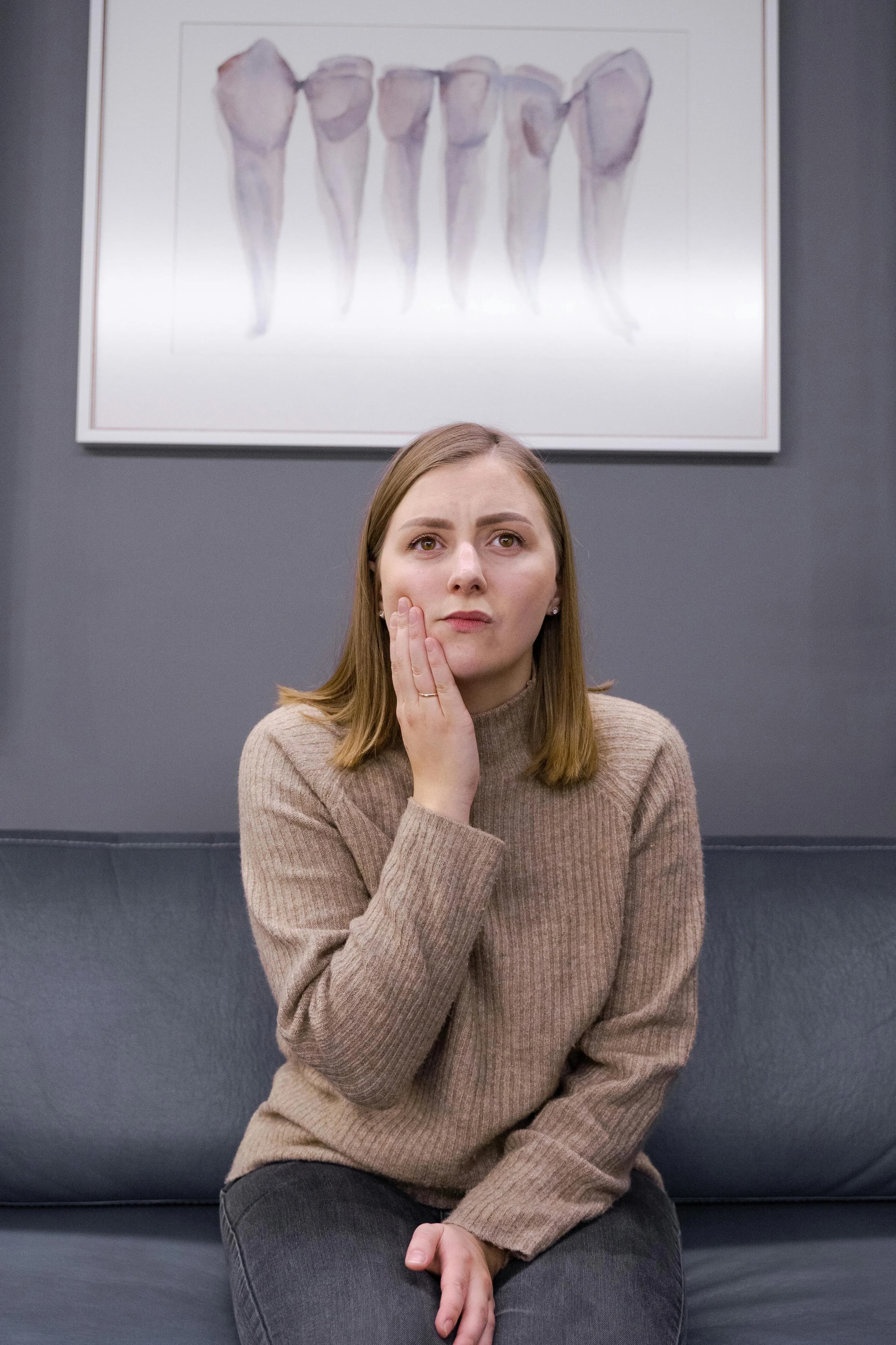 Woman sitting in dental office holding her cheek in pain possibly needing a root canal Astoria NY with tooth diagram on wall.