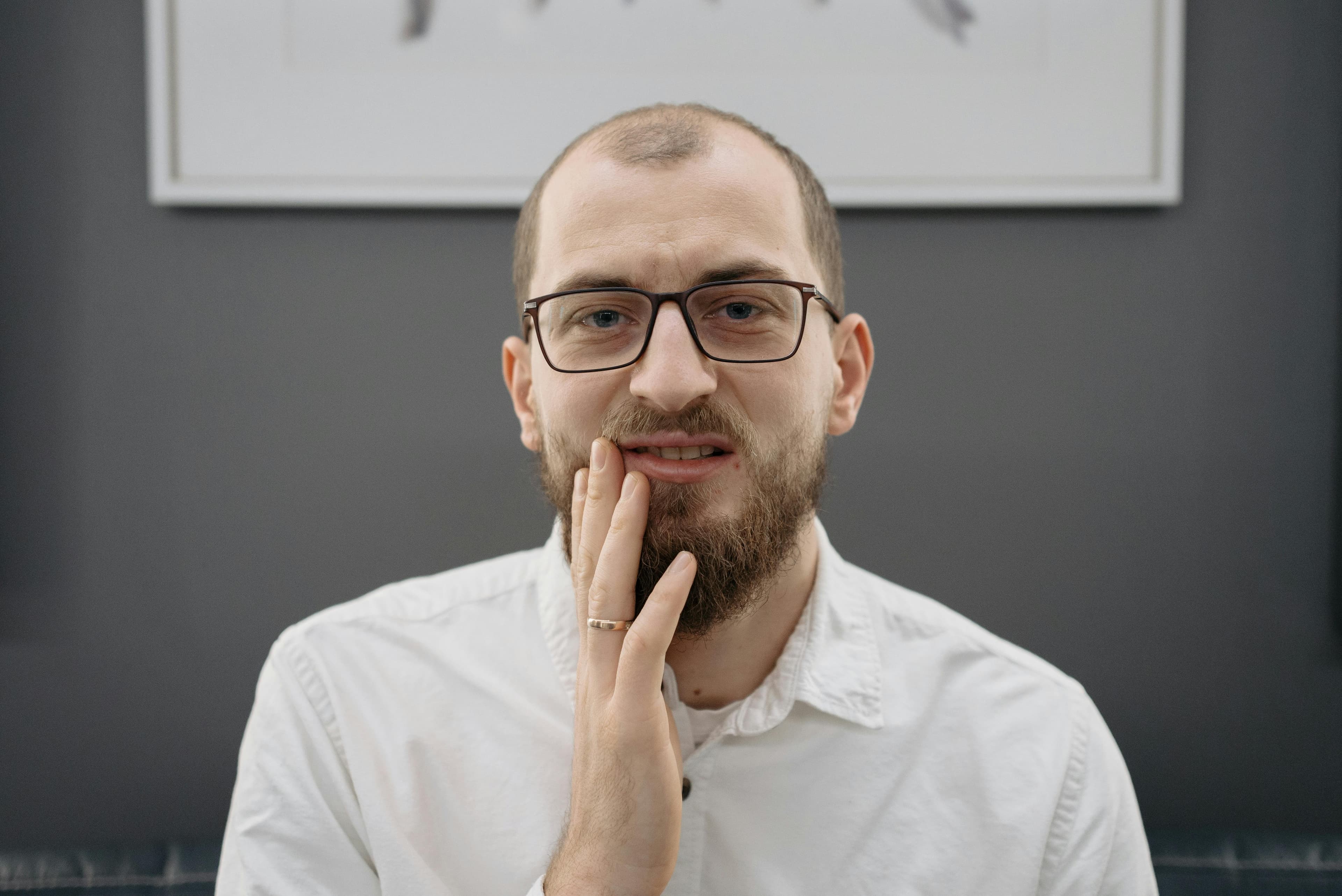 Man With Glasses And Beard Touching Cheek Wearing White Shirt Against Gray Background