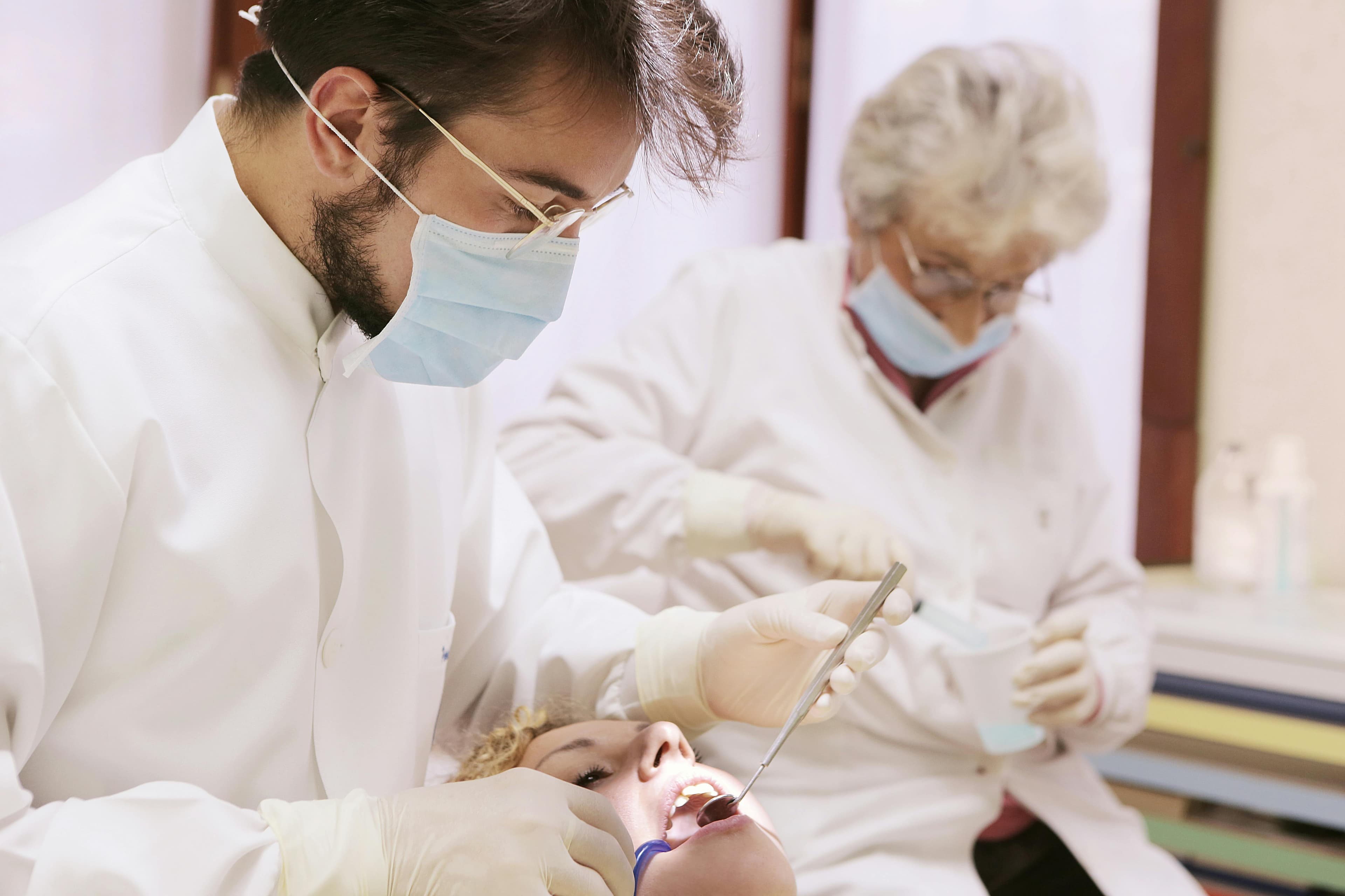 Photo Dentist Wearing Mask And Gloves Examining Patient Mouth With Mirror While Assistant Prepares Tools Demonstrating Dental Emergency Tips In Clinic Setting