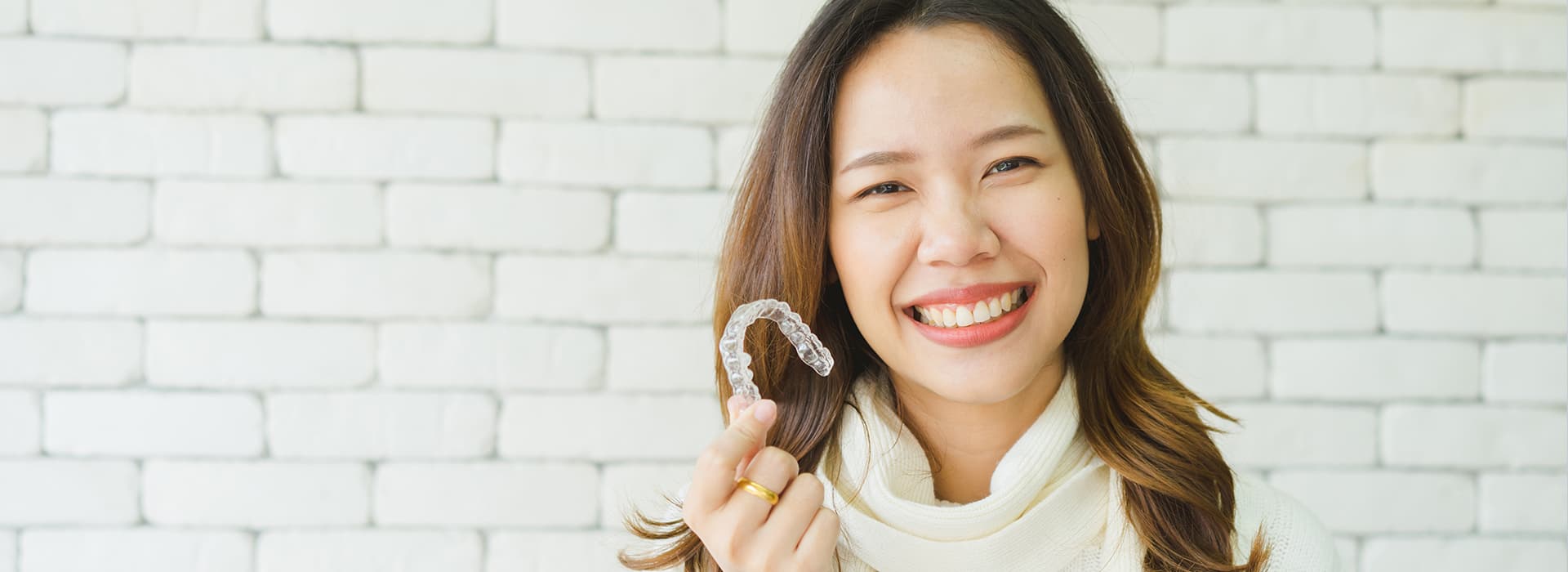 Photo Smiling Woman Holding Clear Aligner Showing Benefits Of Invisalign Against White Brick Wall