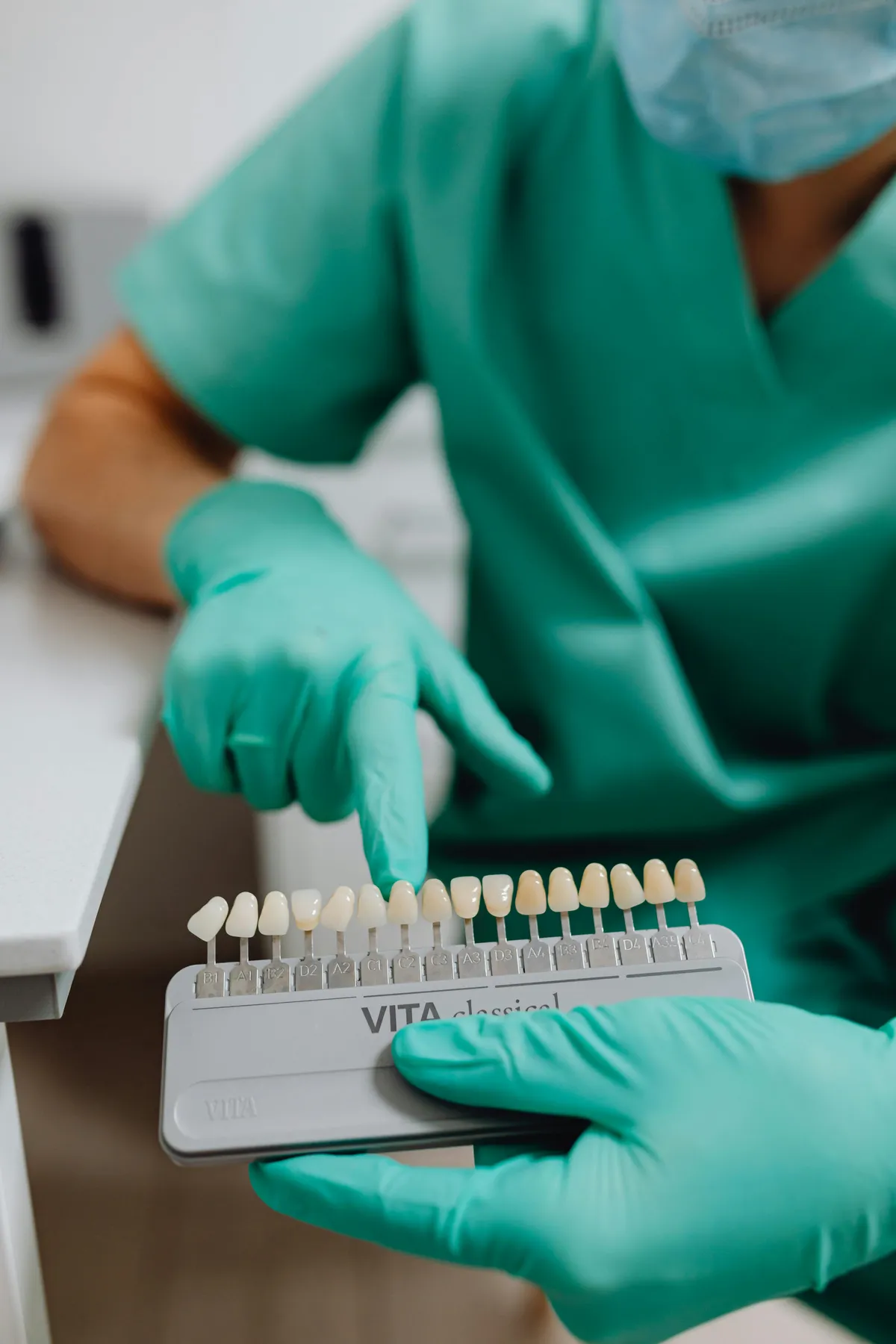 Dentist In Green Scrubs And Gloves Holding Tooth Shade Guide And Pointing At Color Sample To Discuss Can Veneers Be Whitened During Consultation