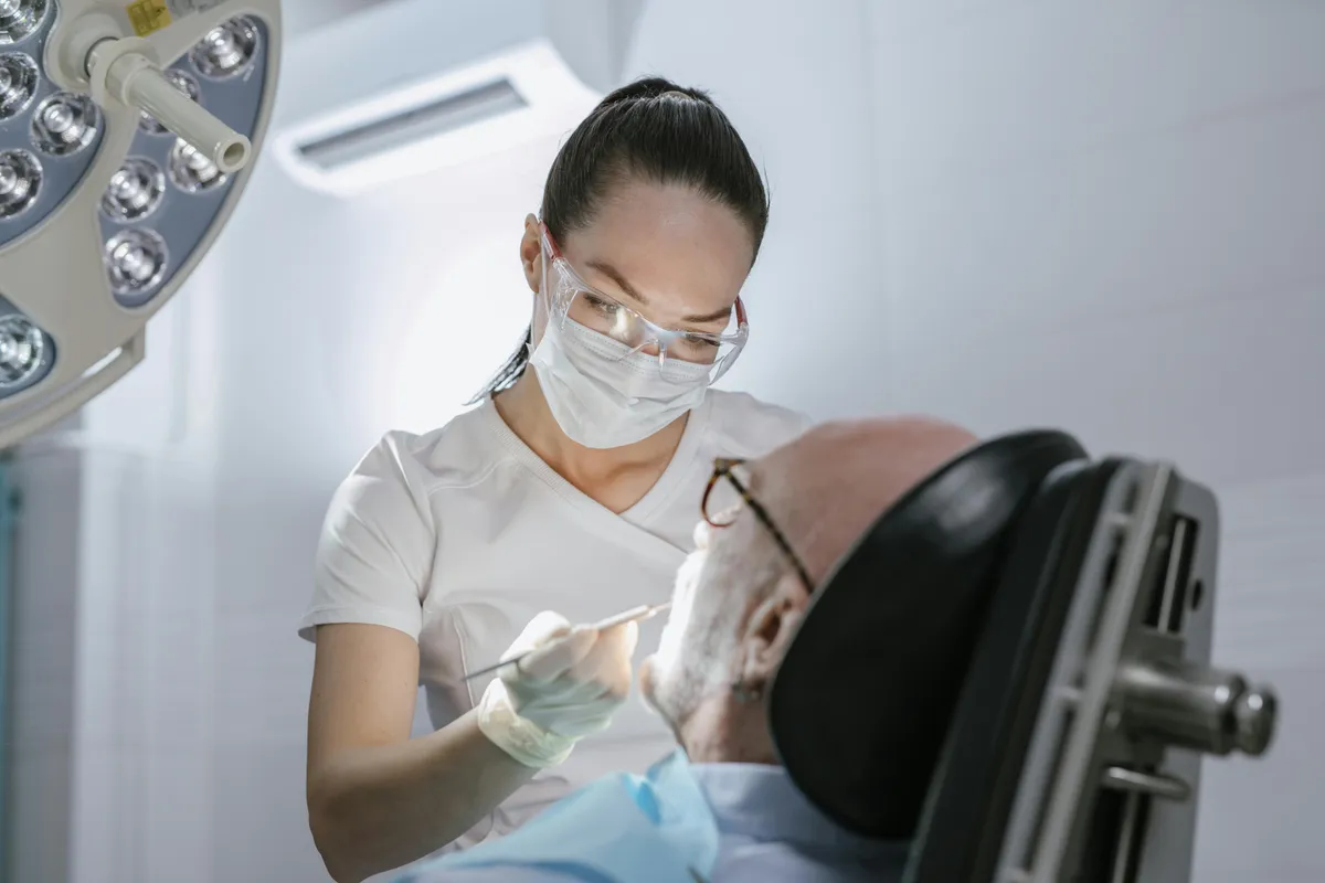 Female Dentist Wearing Mask And Gloves Treating Older Male Patient In Dental Chair Under Surgical Light Discussing How Long Do Bone Grafts Take To Heal During Consultation (1)