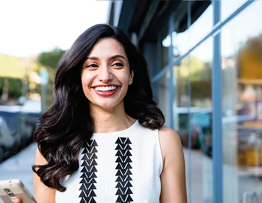 Woman Smiling Outdoors Holding Smartphone Wearing White Top With Black Pattern Standing By Glass Building Showing Confidence And Happiness Related To What Happens After Invisalign
