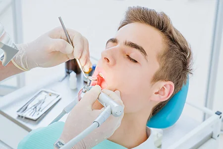 Dentist performing procedure on young male patient using dental tools in clinic with blue chair and tray in background.