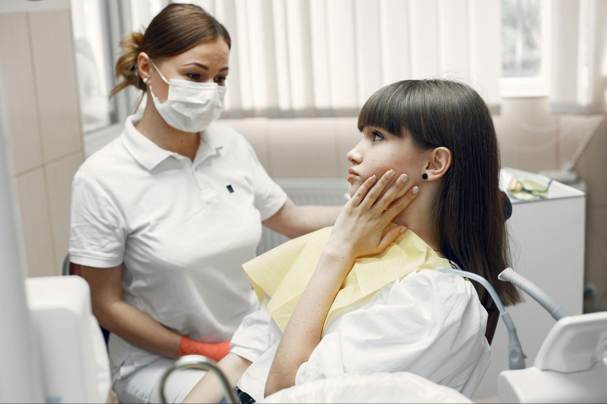 Female dentist wearing mask talking to young patient touching her face in pain during dental consultation in bright clinic room.