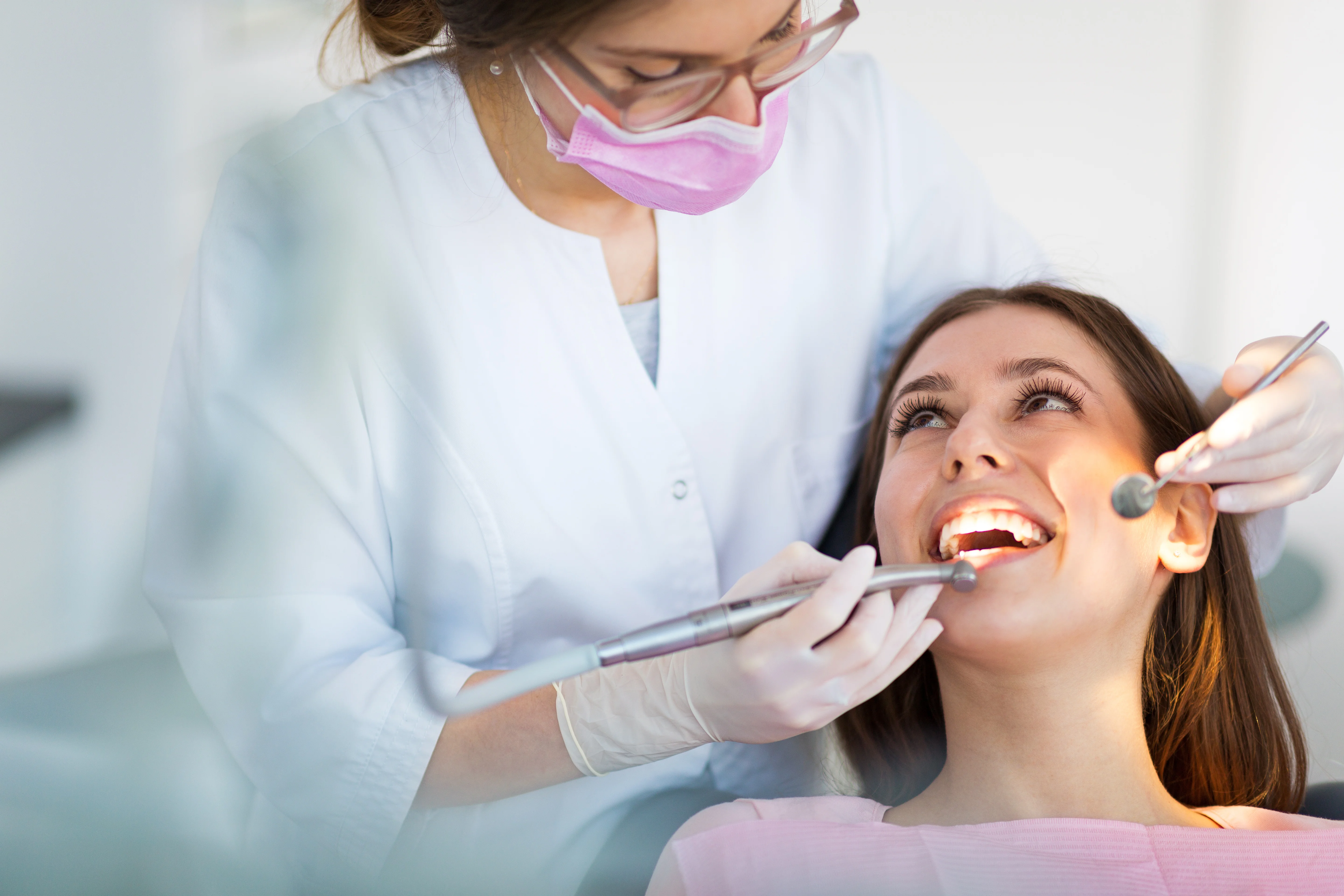 Female dentist wearing pink mask examining smiling female patient using dental tools during oral checkup in modern dental clinic