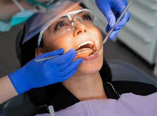 Female patient wearing protective glasses receiving oral exam from dentist in blue gloves using dental mirror and scaler in clinic chair.