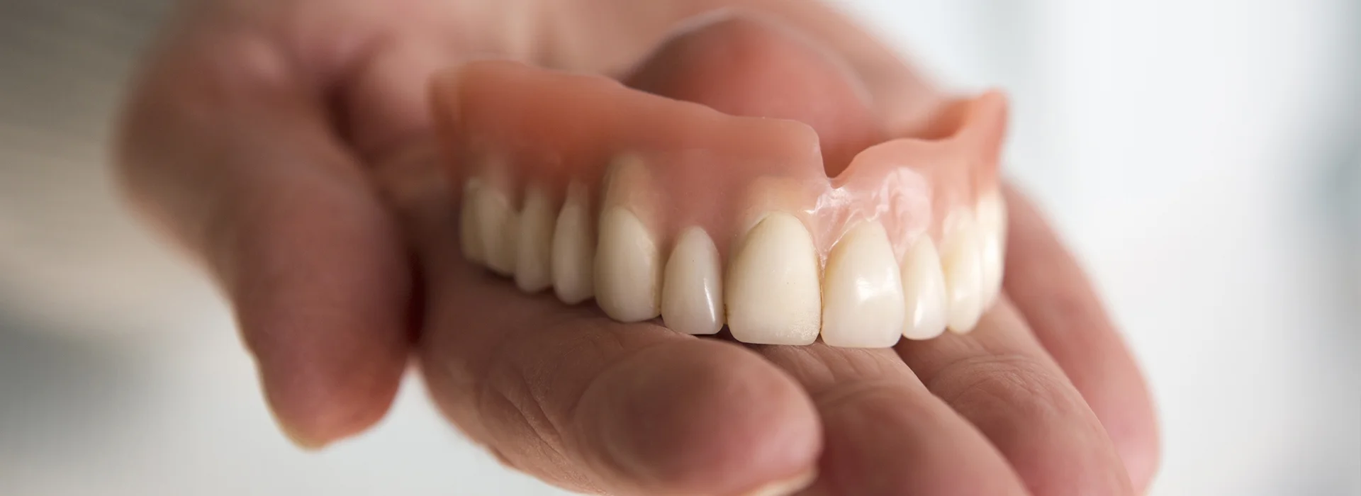 Hand holding upper set of dentures with natural looking teeth in focus against soft blurred background.