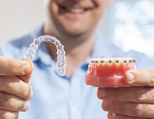 Man holding clear aligner in one hand and dental model with metal braces in other hand demonstrating is Invisalign cheaper than braces decision.