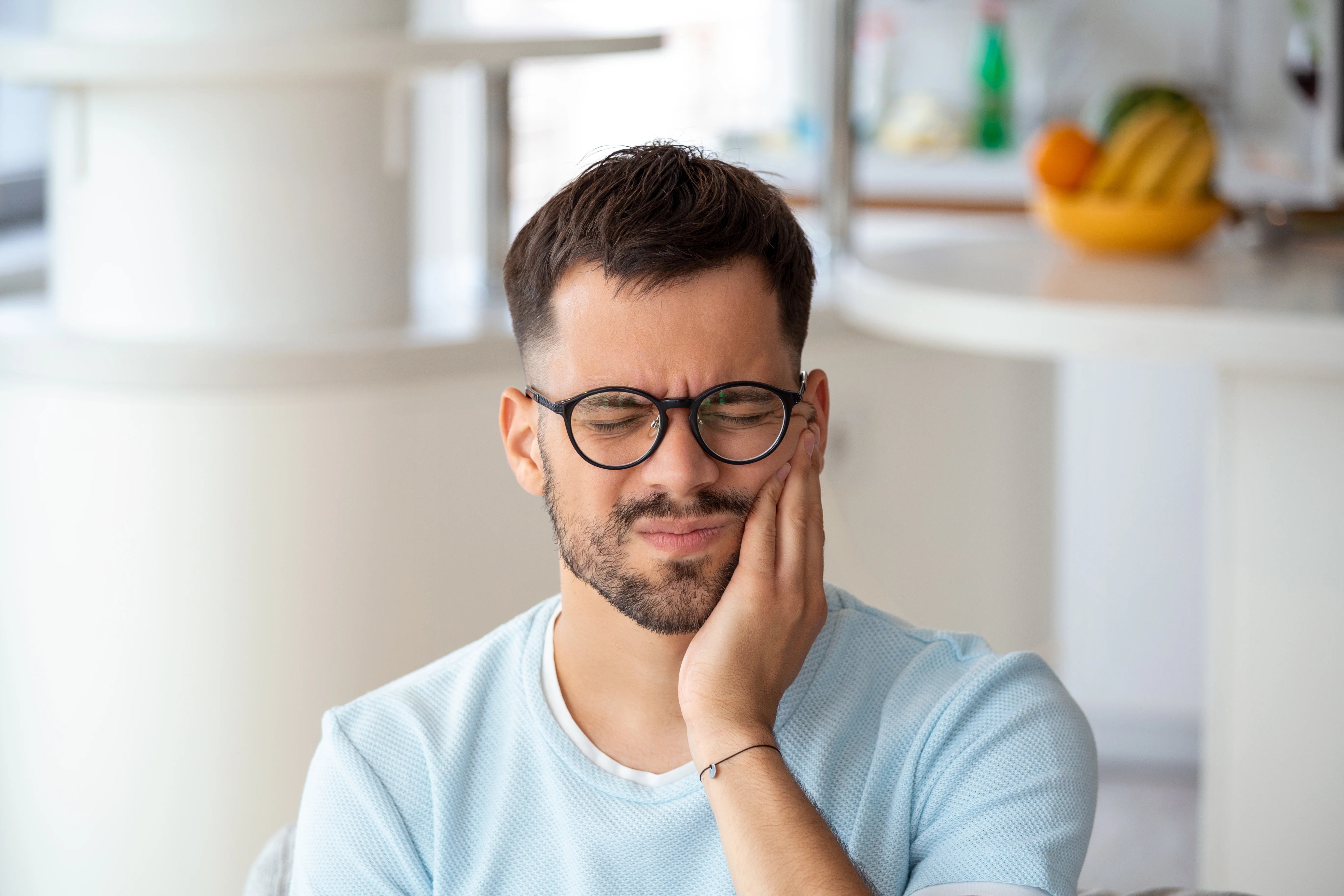 Man holding his jaw in pain while sitting at home wearing glasses and blue shirt expressing discomfort related to disadvantages of mini dental implants.