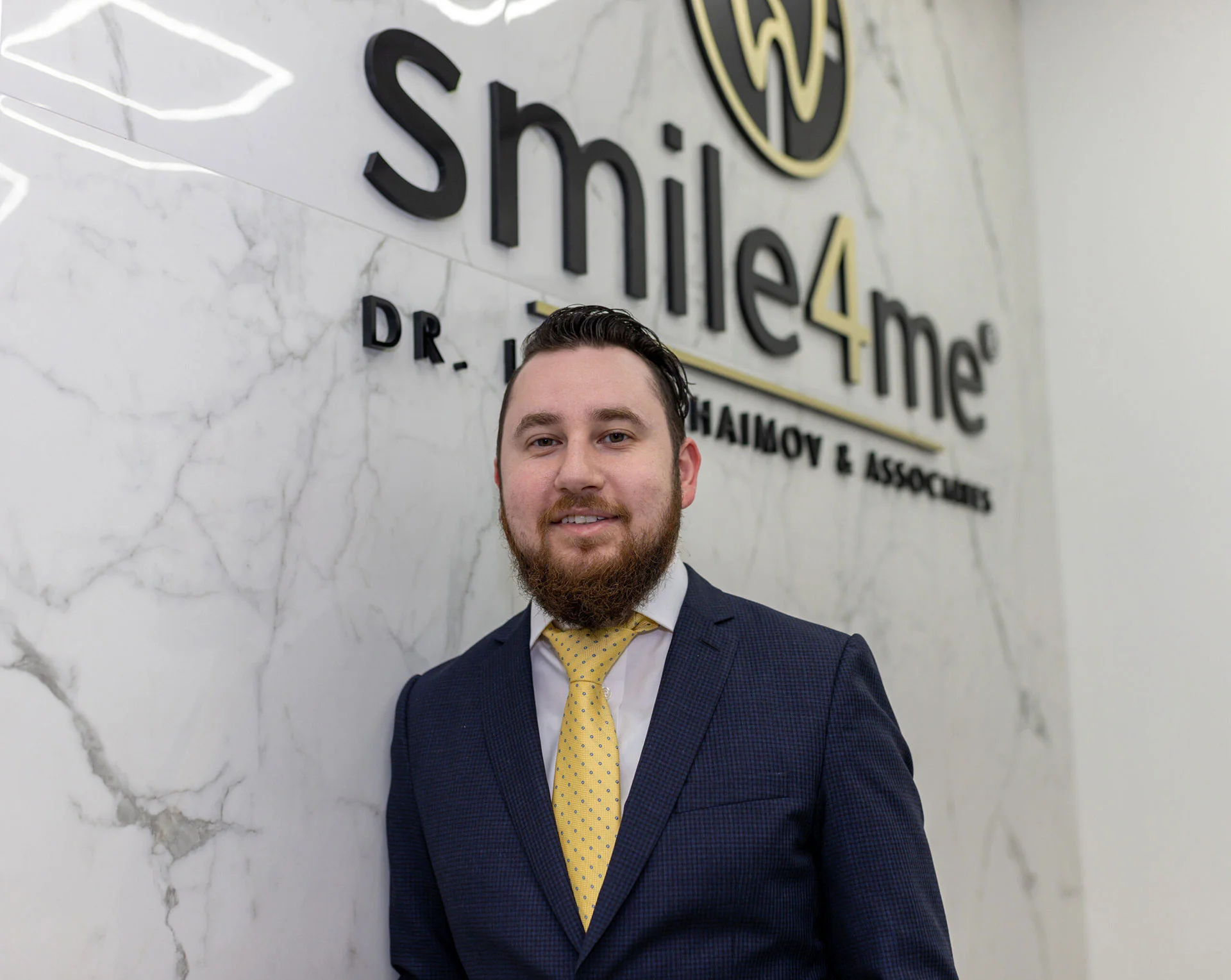 Man in navy suit and yellow tie standing in front of Smile4Me sign on marble wall at dental office.