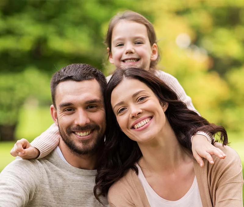 Mother and father with daughter above them outdoors in a park on a sunny day smiling and hugging.