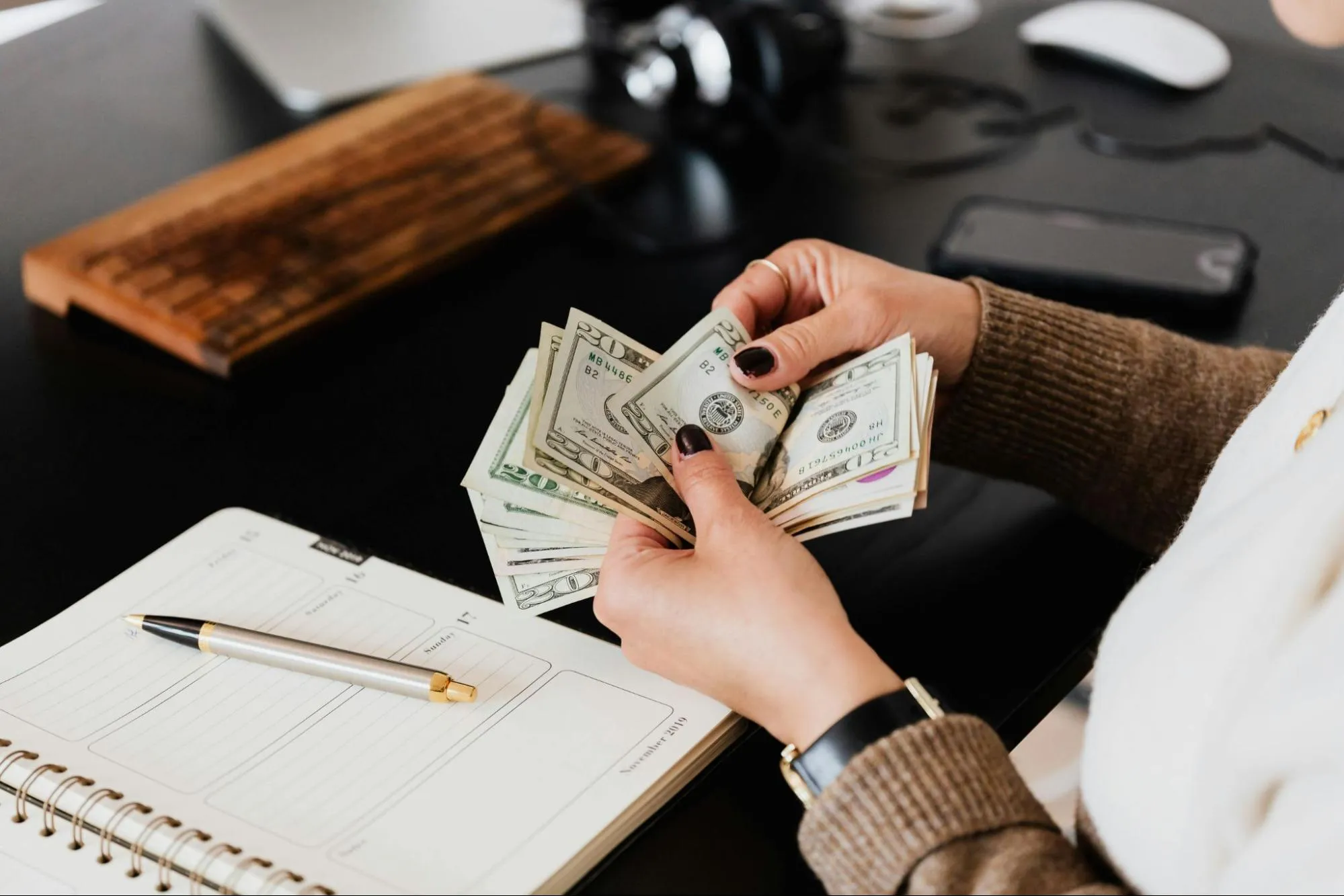 Person counting dollar bills over desk with notebook, pen, smartphone, keyboard, and computer mouse in view.