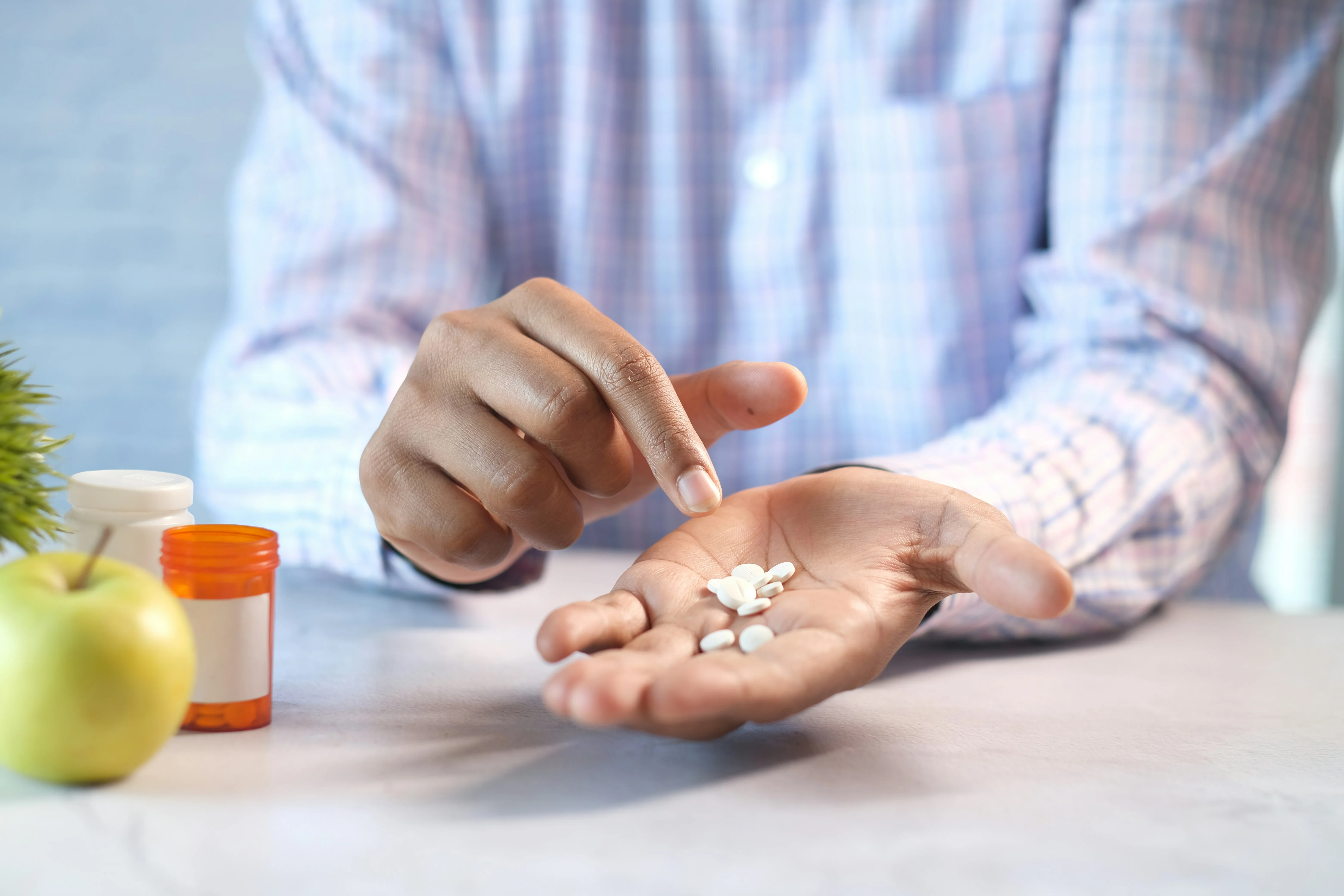 Person in plaid shirt holding white pills in one hand while pointing with other hand with apple and medicine bottles on table.