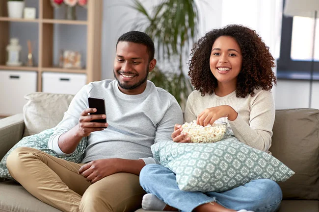 Smiling couple sitting on couch eating popcorn while man holds smartphone in cozy living room with pillows and houseplants in background.