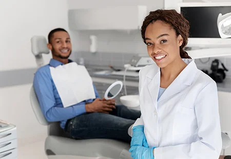 Smiling dentist wearing white coat and blue gloves standing in a modern dental office with a male patient sitting in a dental chair holding a mirror in background.