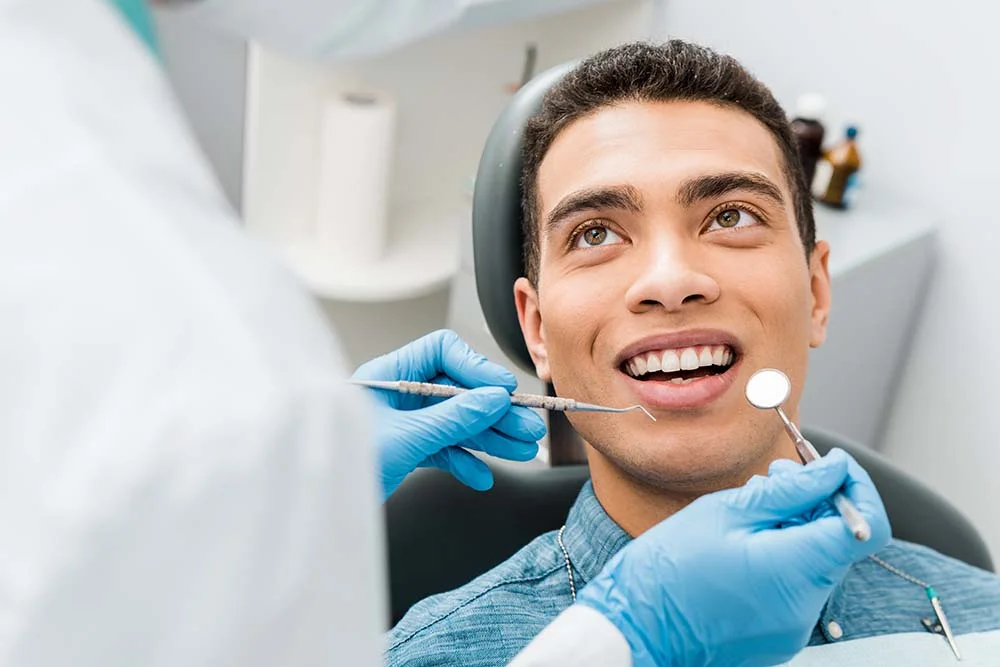 Smiling man sitting in dental chair looking at dentist holding dental tools wearing blue gloves during oral checkup in modern clinic.