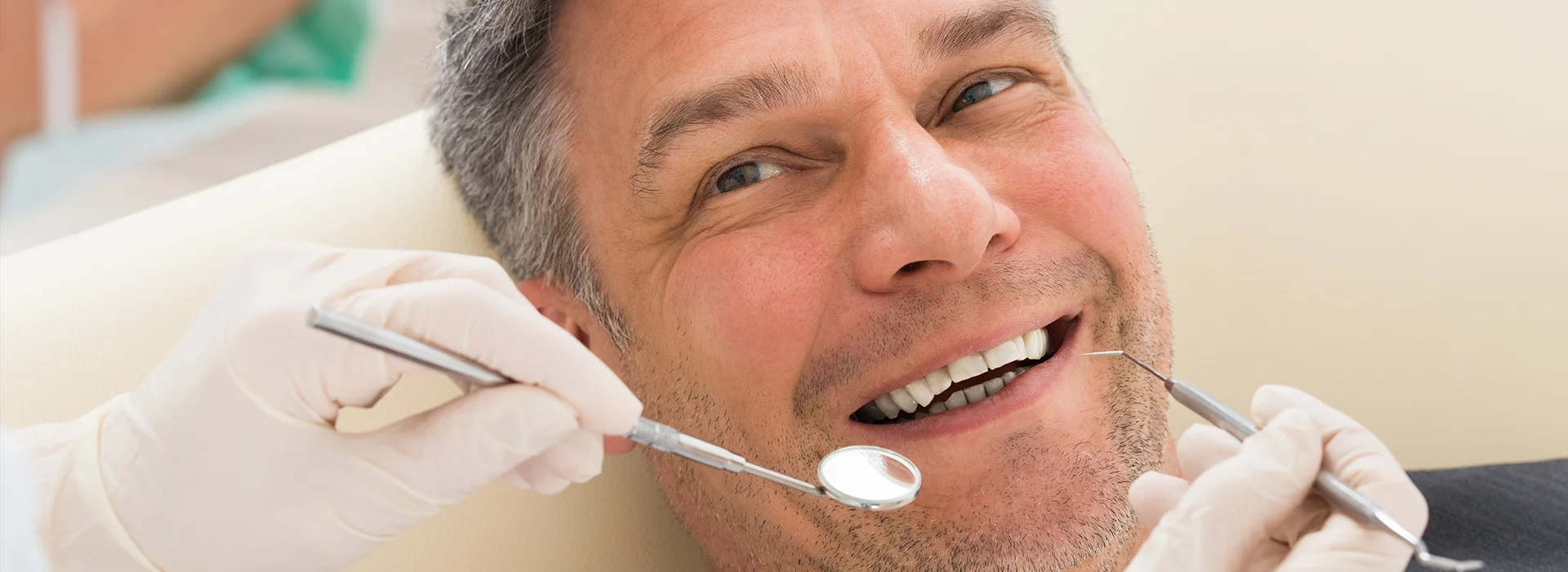 Smiling middle aged man in dental chair during exam with tools near mouth discussing how much do dental implants cost for restoring missing teeth.
              