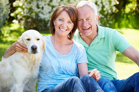 Smiling senior couple sitting on grass with golden retriever outdoors on a sunny day with trees in background.