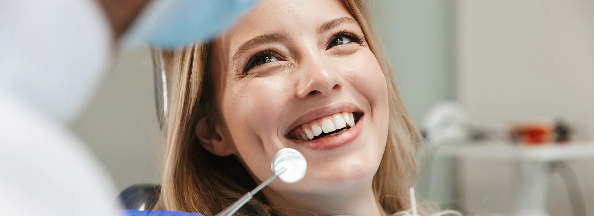 Smiling woman at dental appointment sitting in chair while dentist holds dental mirror for examination in modern clinic.