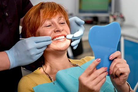 Smiling woman in dental chair holding blue mirror while dentist examines her teeth holding both of his hands with blue gloves to her mouth.