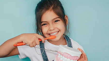 What is preventive dentistry smiling young girl holding toothbrush and large dental model practicing proper toothbrushing technique against blue background.