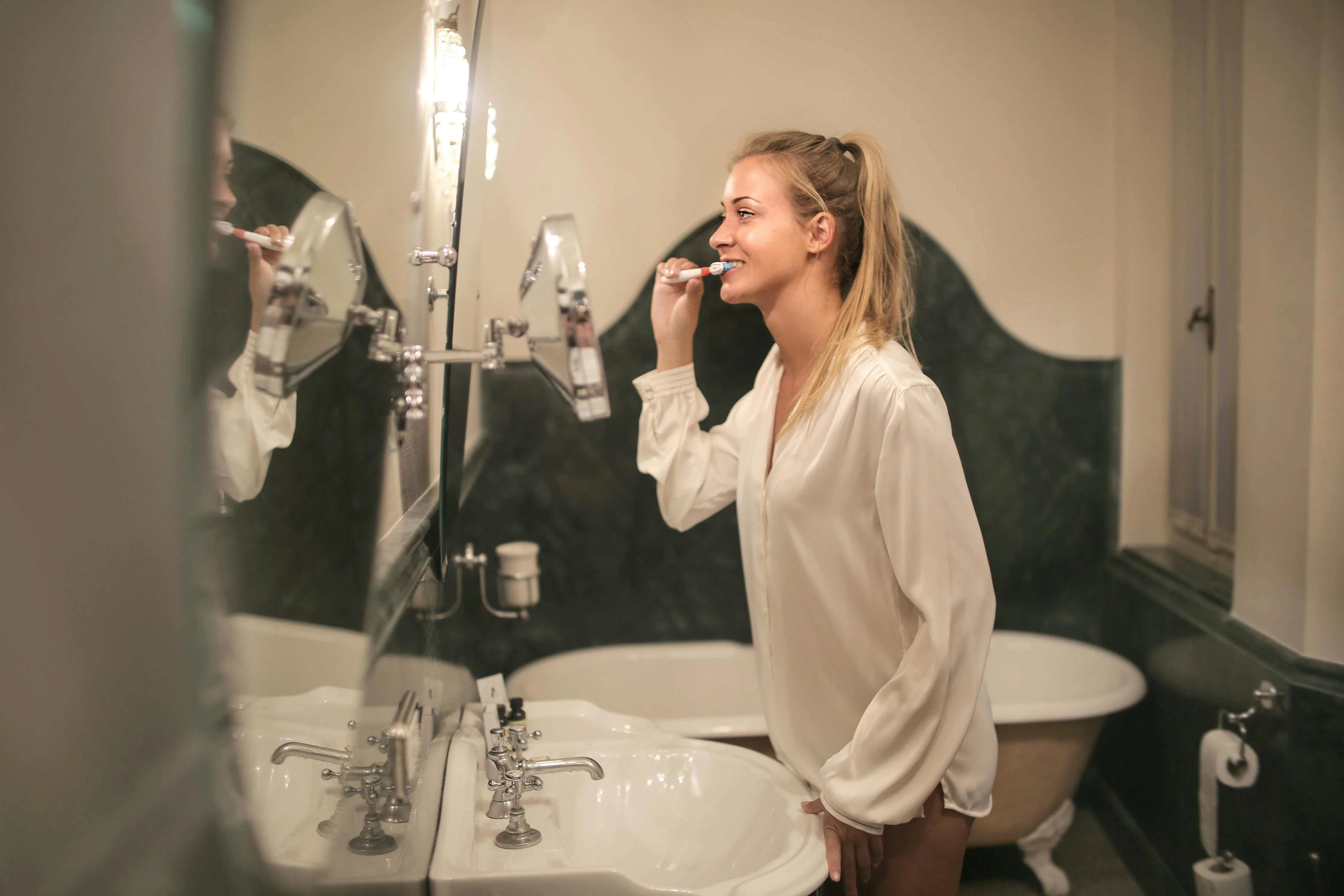 Woman brushing teeth in bathroom while smiling and looking into mirror wearing white long sleeve shirt standing by sink.