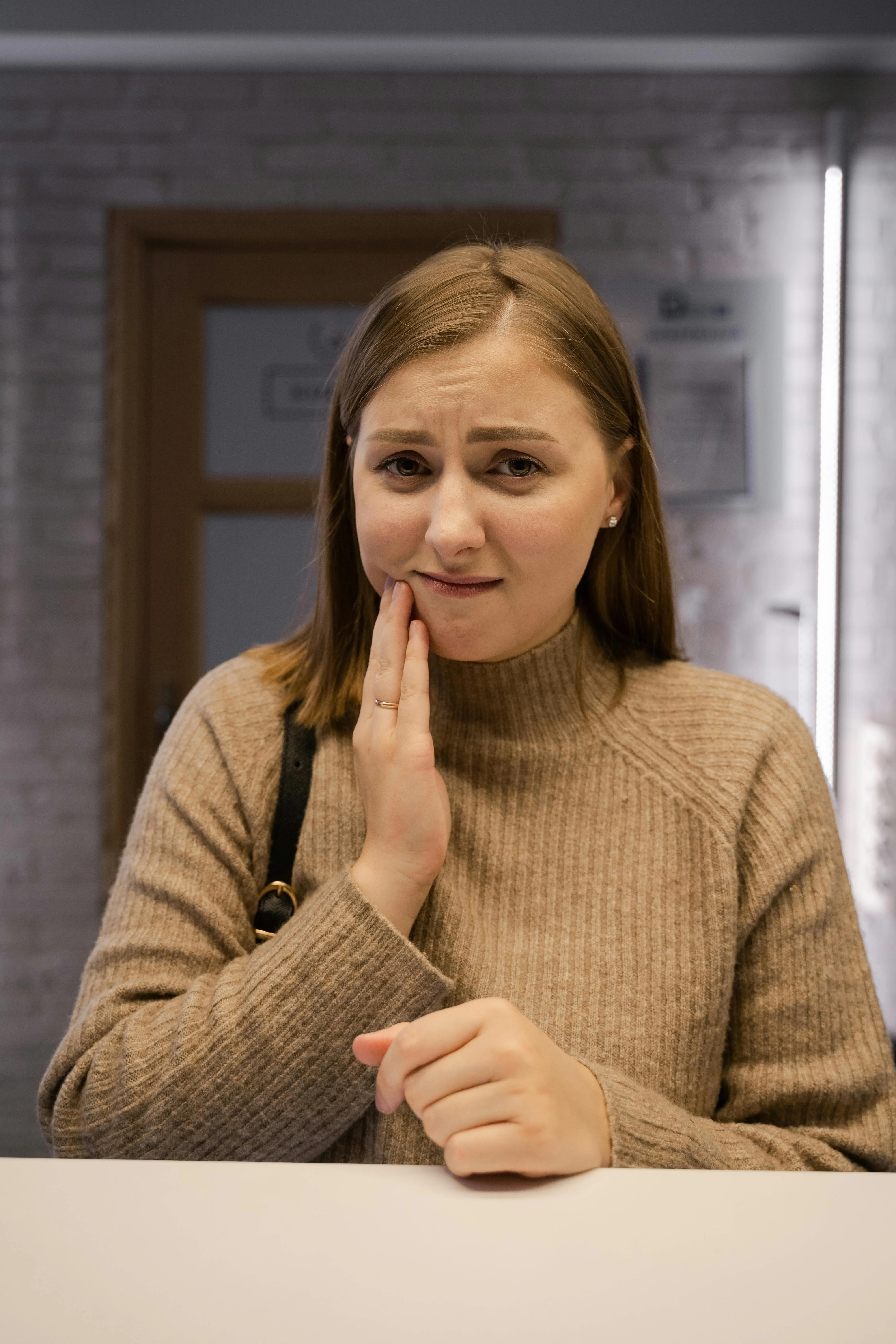 Woman holding her jaw in pain standing at reception desk showing visible discomfort as one of the dental emergency examples due to severe toothache.