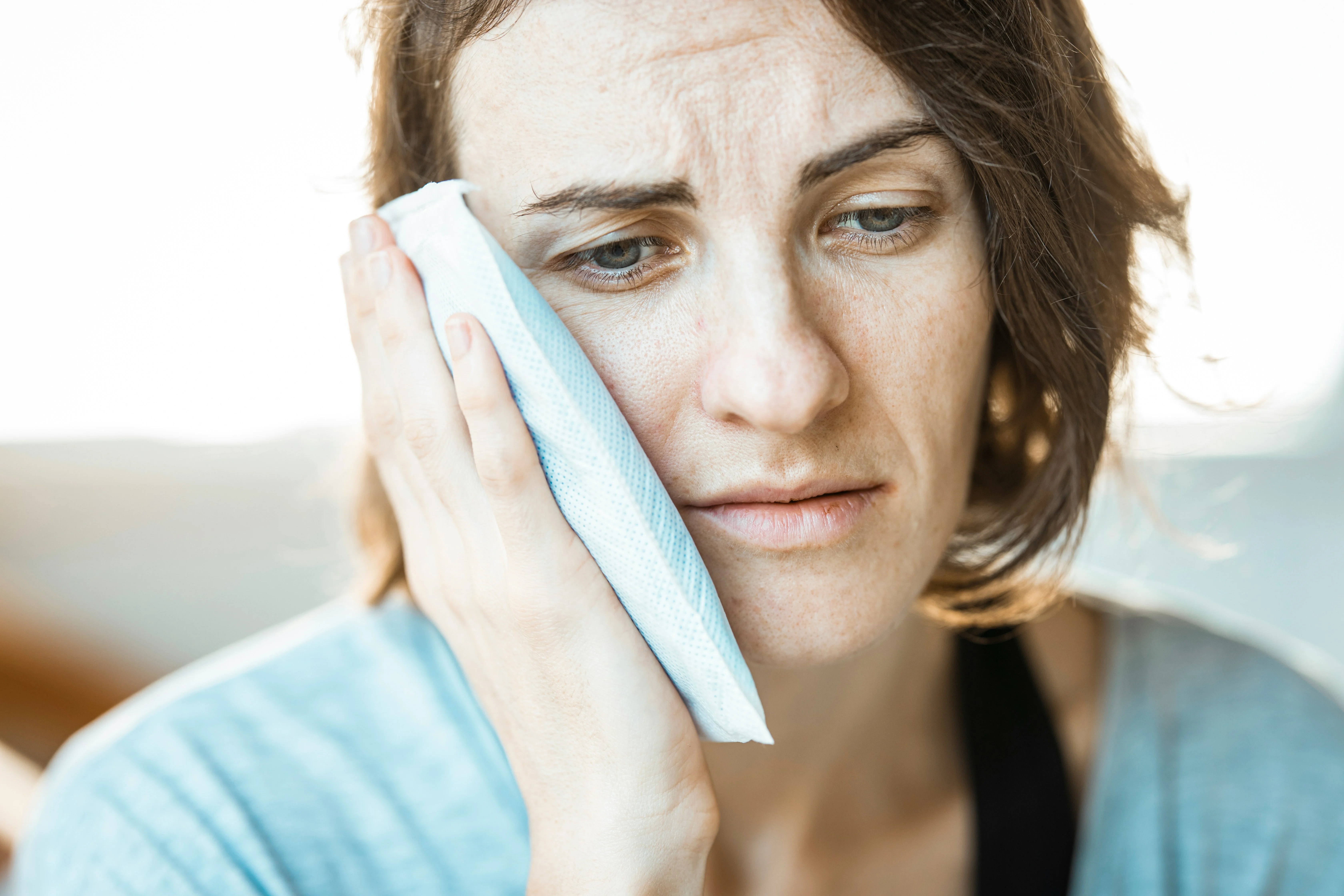 Woman holding ice pack to cheek looking uncomfortable indoors illustrating situation where you wonder can you go to the ER for tooth pain.