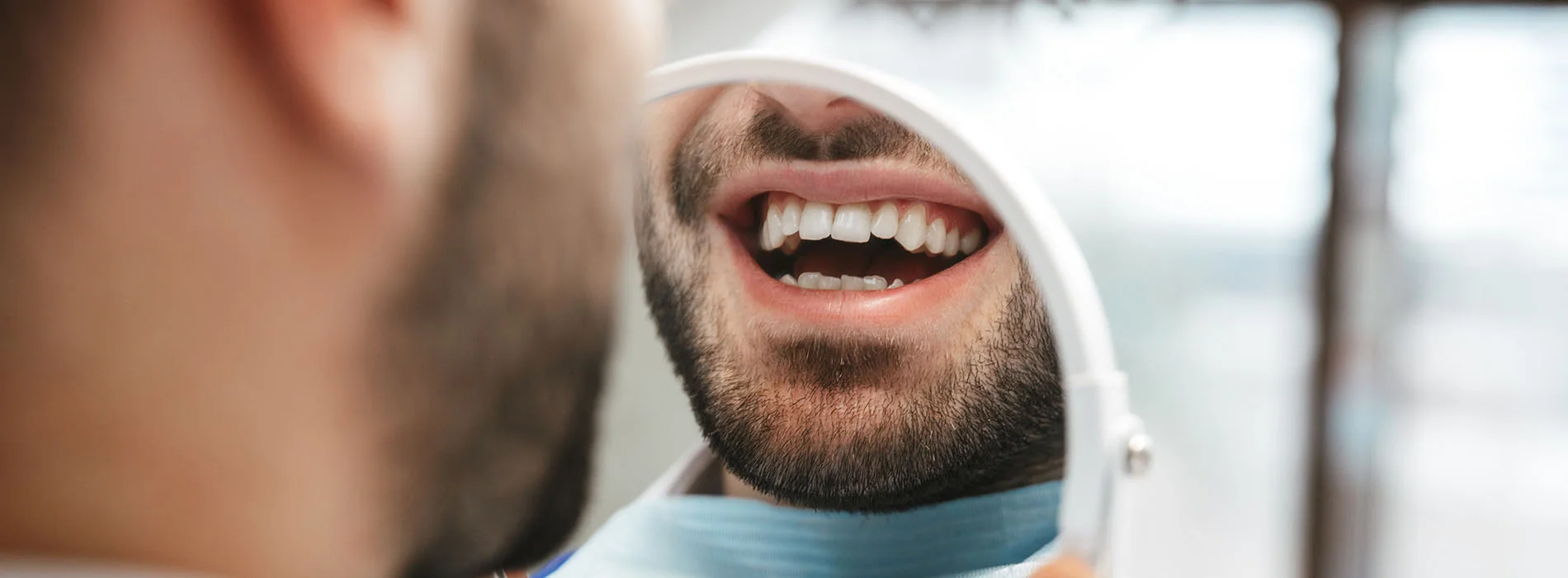 Close up of man smiling while looking at his teeth in a handheld mirror during dental checkup in a bright office setting.