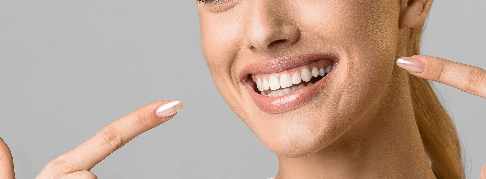 Close up of smiling woman pointing at her white teeth showing results of cosmetic dentistry treatment with polished nails and neutral background.