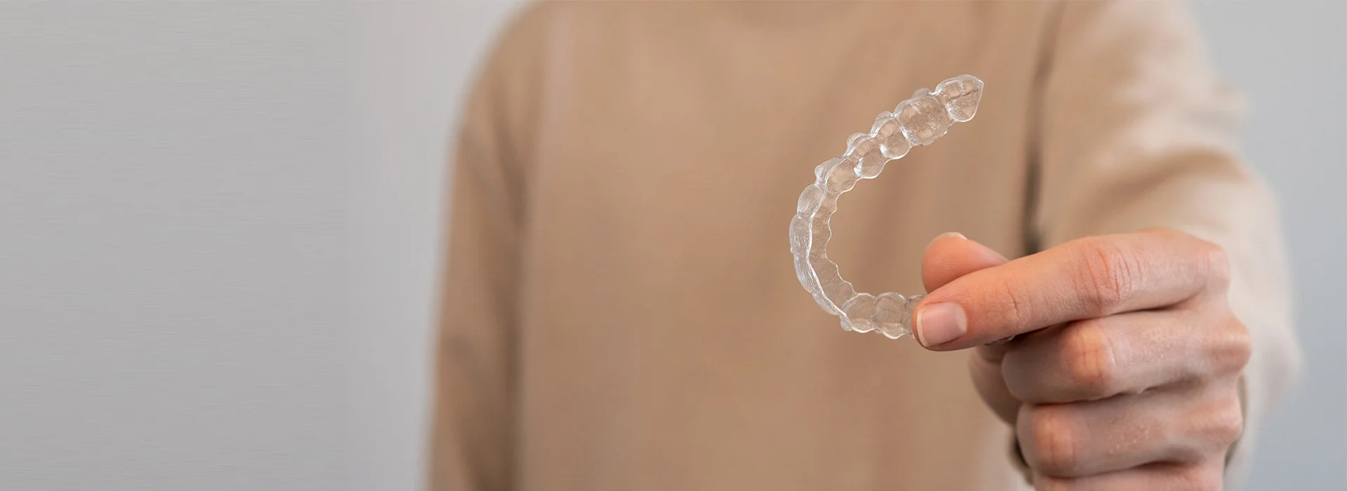 Hand holding clear Invisalign aligner close to camera against neutral background raising question is Invisalign worth it for straightening teeth discreetly.
