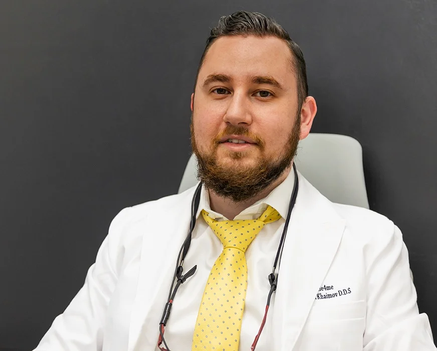 Man wearing white coat and yellow tie sitting in chair with glasses on strap around neck against dark background looking directly at camera.