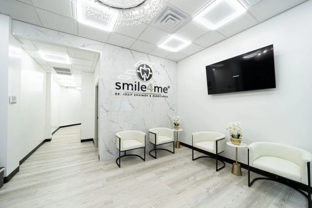 Modern dental office waiting room with white chairs, marble wall, Smile4Me logo, TV screen, and decorative flowers on small gold accented tables.