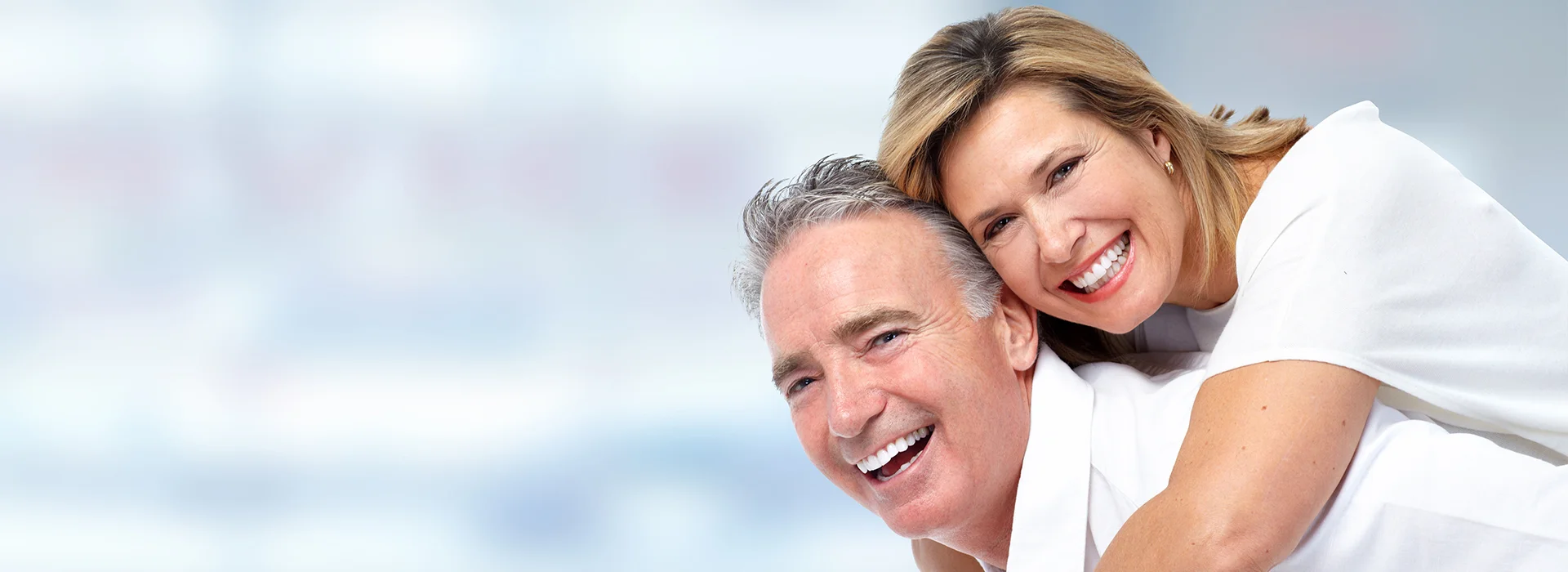 Smiling middle aged couple in white clothing hugging and laughing while posing together in front of light blue background.