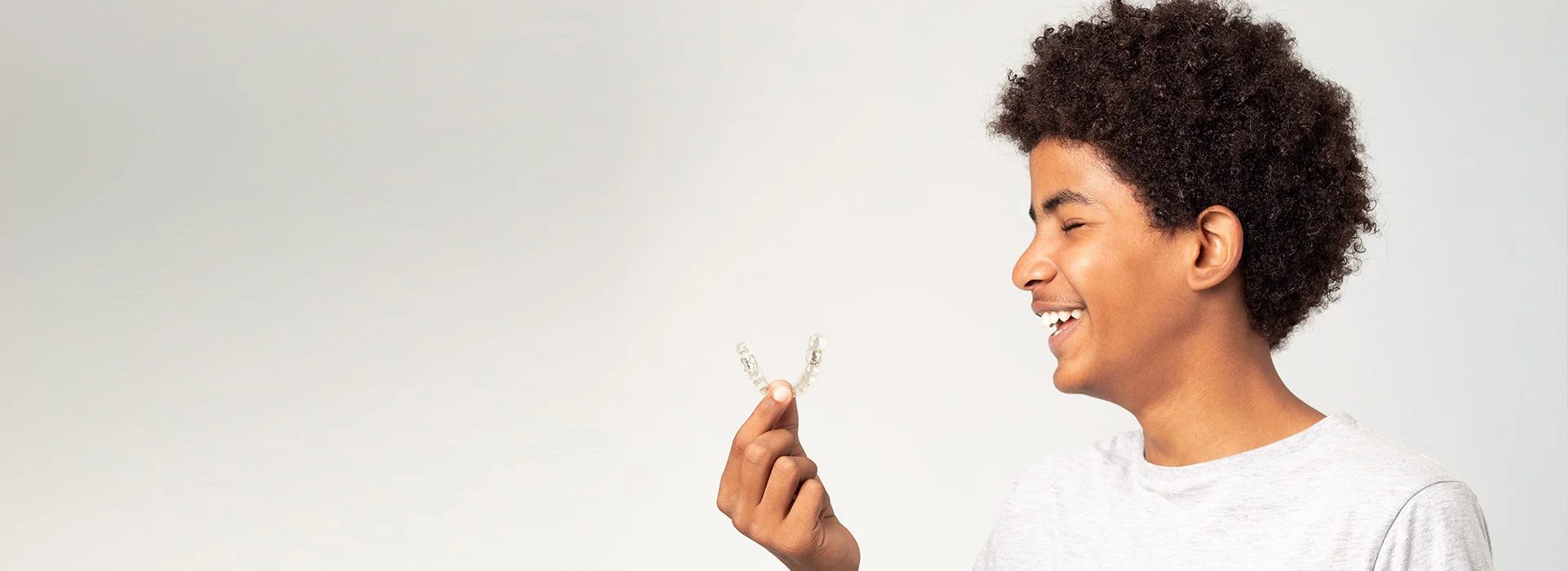 Smiling teen boy holding clear aligner in hand wearing white shirt against neutral background.