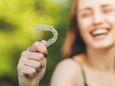 Smiling woman outdoors in natural light holding clear aligner in focus with green blurred background displaying Invisalign tray.