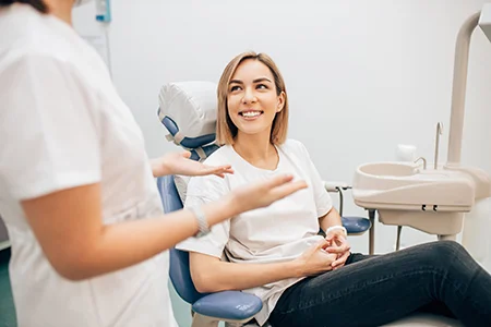 Smiling woman sitting in dental chair listening to dentist in white uniform inside clean modern clinic with sink and dental equipment in background.