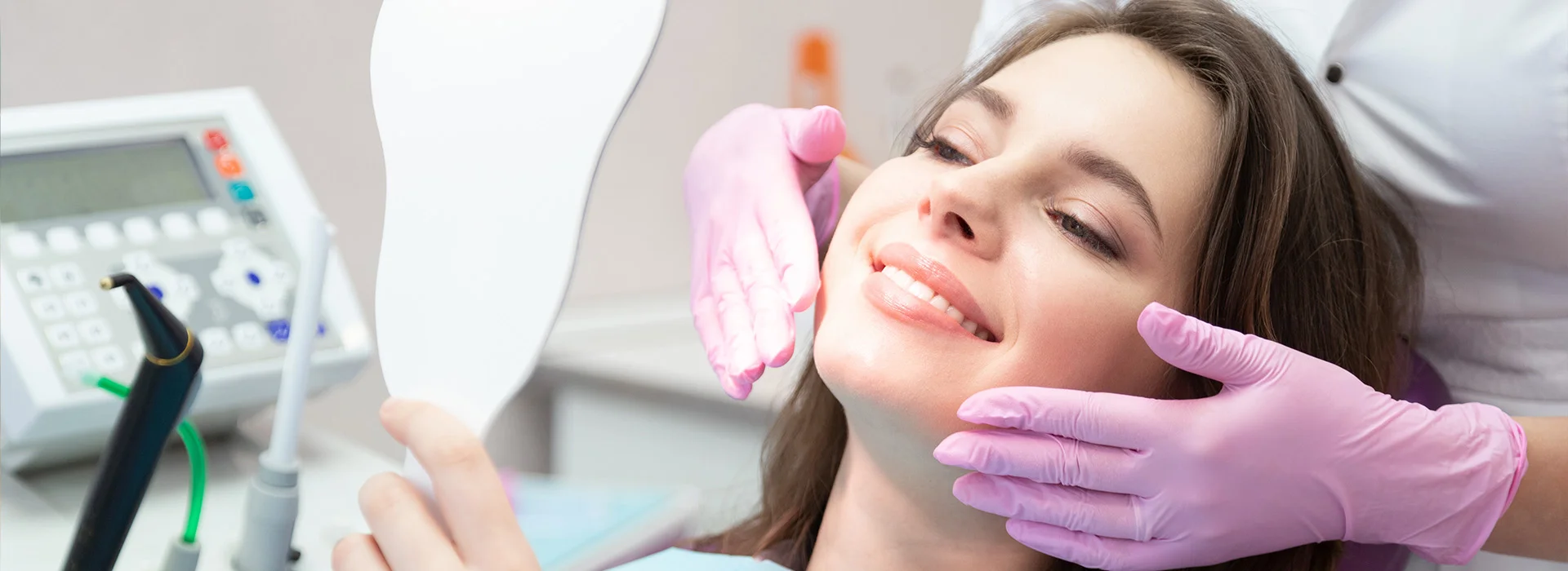 Woman smiling in dental chair holding mirror while dentist in pink gloves checks results of her smile makeover in modern dental office setting.